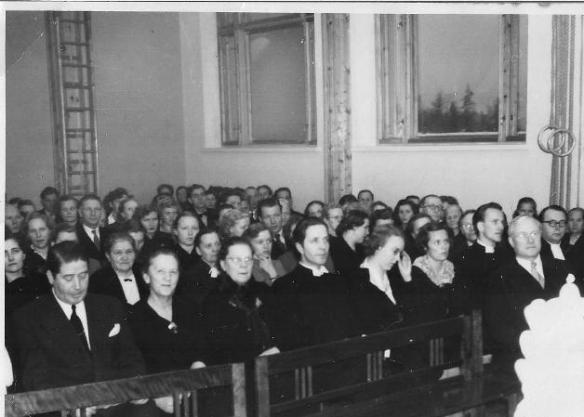 Kirkon 25-vuotisjuhlien päiväjuhlassa yhtiön teknillinen johtaja Väinö Huida on yksin etupenkissä. Toisella rivillä mm Hannikainen, silloinen väliaikainen pastori Lappalainen ja tuleva kirkkoherra Nuottamo vaimoineen.